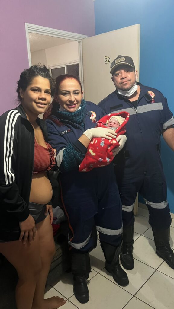 A fotografia captura um momento de celebração e cuidado, com uma mãe sorrindo ao lado de dois profissionais de saúde que seguram um recém-nascido envolto em uma manta vermelha estampada. A composição da imagem foca nas pessoas em primeiro plano, com a mãe posicionada à esquerda e os dois profissionais à direita, criando uma linha horizontal que atrai o olhar. A cena parece ter sido capturada logo após o nascimento, com a mãe demonstrando alívio e felicidade, e os profissionais exibindo um semblante de orgulho e profissionalismo. A presença do recém-nascido, o centro da atenção, adiciona um elemento de ternura e importância ao quadro. O enquadramento médio-close destaca as expressões faciais e a interação entre os indivíduos. A mãe é uma mulher jovem, com pele morena, cabelo escuro preso em um coque bagunçado e um sorriso radiante. Ela veste uma blusa de top vermelho e uma jaqueta preta com listras brancas nas mangas, revelando uma barriga de gravidez recente. Ao seu lado, uma mulher com cabelo ruivo e liso, vestindo um uniforme azul escuro com detalhes em verde e um broche identificador, segura o bebê com luvas brancas. O bebê, envolto em uma manta vermelha com estampa de estrelas e desenhos animados, tem um gorro branco na cabeça e parece adormecido. Ao lado dela, um homem com boné, também em uniforme azul escuro com detalhes prateados, observa a cena com uma expressão serena, usando uma máscara de proteção. A fotografia é um registro realista, possivelmente tirada com uma câmera digital ou de smartphone, sem filtros artísticos elaborados. O estilo é documental, com ênfase na captura genuína do momento. A iluminação parece ser artificial e direta, vinda de cima, o que cria algumas sombras suaves no fundo. A escolha de cores é natural, sem saturação excessiva, transmitindo a autenticidade da cena. O cenário é um ambiente interno simples, possivelmente um quarto de hospital ou uma casa, com paredes pintadas em tons de lilás e azul claro. Uma porta branca, com o número "22" visível, está aberta ao fundo, sugerindo um espaço de transição. O piso é de azulejo branco quadriculado. A iluminação interna, embora não espetacular, é suficiente para iluminar os rostos e destacar os sujeitos principais, contribuindo para uma atmosfera acolhedora e de realização.