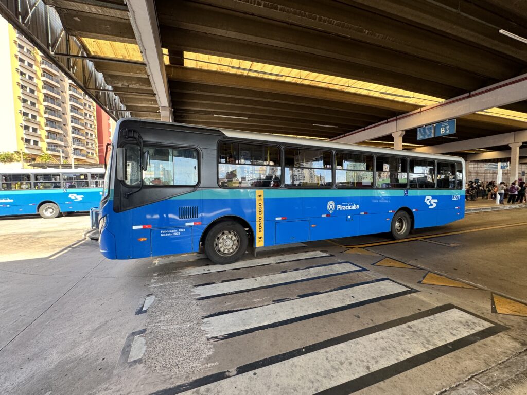 A imagem mostra um ônibus do transporte coletivo urbano de Piracicaba estacionado em uma plataforma de terminal de integração. O veículo é predominantemente azul, com detalhes em preto na parte frontal e superior, e traz a identificação da Prefeitura de Piracicaba e da empresa operadora na lateral. Próximo à porta dianteira há uma faixa amarela indicando um ponto cego, com ícones de atenção para pedestres e ciclistas.