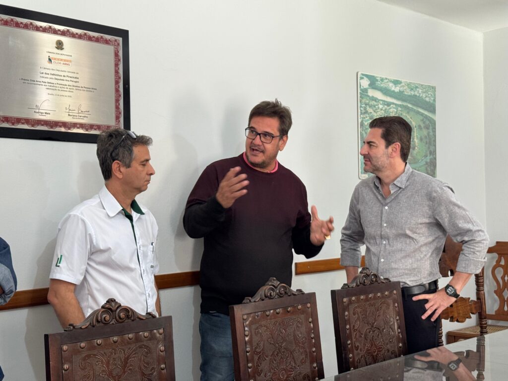 A imagem mostra três homens em uma sala, em uma conversa aparentemente profissional ou de negócios. * O homem à esquerda veste uma camisa social branca com detalhes em verde. * O homem ao centro, que parece conduzir a conversa com gestos, usa um suéter de gola careca na cor vinho sobre uma camisa rosa e óculos de grau. * O homem à direita veste uma camisa social cinza de mangas dobradas e relógio de pulso. Eles estão posicionados em frente a uma mesa de madeira com cadeiras de design clássico e entalhadas. Na parede, à esquerda, há um quadro com um diploma ou certificado emoldurado da Câmara dos Deputados, concedido ao "Lar dos Velhinhos de Piracicaba". À direita, há um quadro com uma foto aérea de uma paisagem, possivelmente uma região com rio e vegetação. O ambiente é iluminado e sugere um espaço administrativo ou de reuniões.