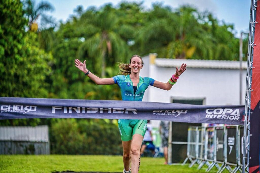 A imagem mostra uma atleta feminina cruzando a linha de chegada de uma competição esportiva ao ar livre. Ela está vestindo um traje esportivo nas cores verde e azul com o logo "care club" no peito. A atleta está sorrindo e levantando os braços em sinal de vitória, segurando a fita da linha de chegada que tem a palavra "FINISHER" impressa, além do nome do evento "COPA INTERIOR" e a marca "CHELSO SPORTS & BUSINESS". Ao fundo, há árvores tropicais e vegetação, sugerindo um ambiente quente e ensolarado. A foto transmite uma sensação de conquista e celebração.
