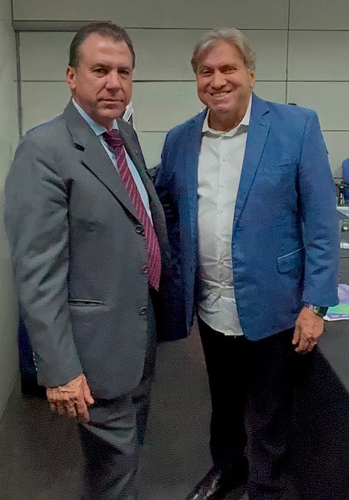 A imagem mostra dois homens em uma foto posada, possivelmente em um ambiente profissional ou de evento. O homem à esquerda veste um terno cinza com gravata listrada, enquanto o da direita está com um paletó azul, camisa branca e calça preta. Ambos estão sorrindo.