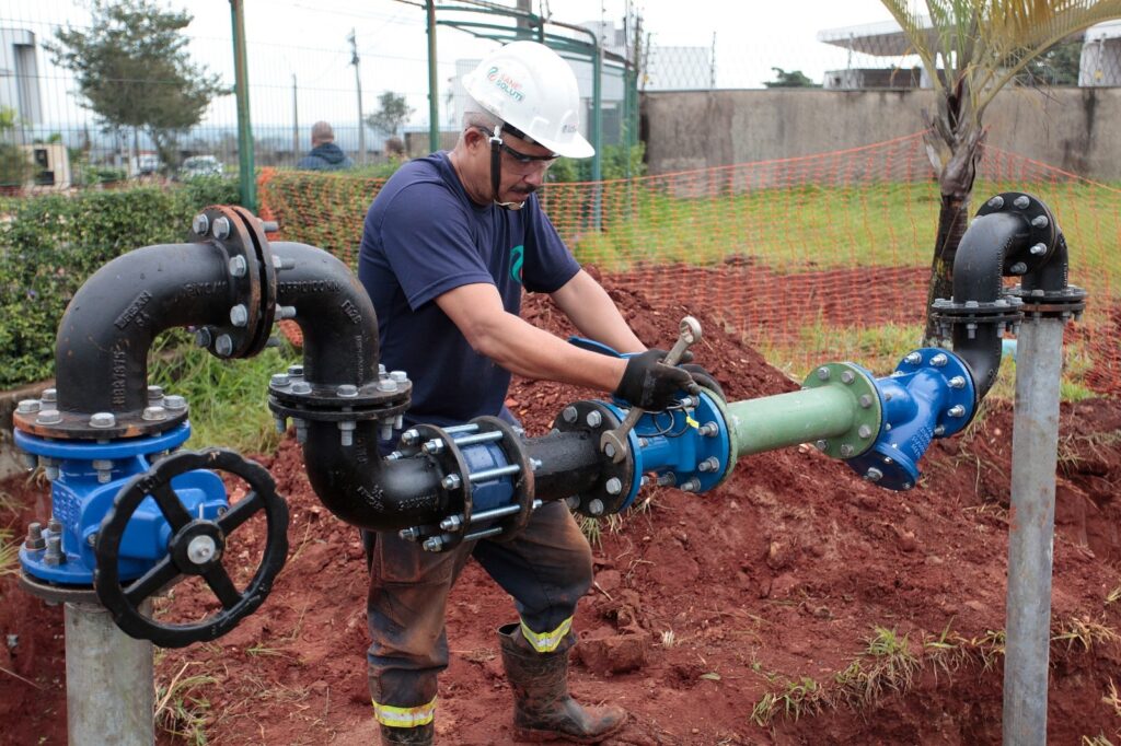A imagem mostra um trabalhador masculino em um ambiente industrial ao ar livre, provavelmente em um canteiro de obras ou local de manutenção. Ele está concentrado em uma tarefa que envolve tubulações industriais, aparafusando ou ajustando uma conexão com uma chave inglesa. O homem veste uma camiseta azul escura, capacete branco de segurança com um logotipo, óculos de proteção e luvas pretas, indicando que está seguindo normas de segurança no trabalho. As tubulações são principalmente pretas com algumas partes azuis, dispostas em formato de “U” e conectadas a um tubo vertical. O solo ao redor é avermelhado e aparenta estar escavado recentemente, sugerindo que o trabalho está em fase de instalação ou reparo. Ao fundo, há uma cerca verde metálica, uma tela laranja de segurança e vegetação esparsa, além de algumas pessoas ao longe. O céu parece nublado, com luz difusa, o que evidencia melhor os detalhes da cena. No canto superior esquerdo da imagem, aparece parcialmente o texto “SANE SOLUTI”, que provavelmente faz parte do nome ou logotipo da empresa responsável pelo serviço. A imagem transmite uma atmosfera de trabalho técnico e especializado em infraestrutura hidráulica ou saneamento básico.