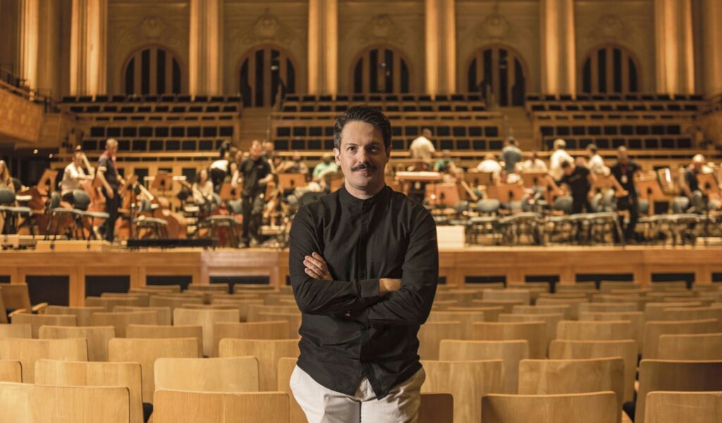 A imagem mostra um homem em plano médio, posicionado no centro de uma sala de concertos vazia. Ele está de braços cruzados, olhando diretamente para a câmera, com uma expressão tranquila e confiante. Ele tem cabelo escuro, bigode, e veste uma camisa preta de botões e calças claras. Ao fundo, no palco, há uma orquestra se preparando para uma apresentação; vários músicos estão visíveis com seus instrumentos, incluindo violoncelos, criando um ambiente de atividade, embora desfocado. A sala de concertos tem uma arquitetura clássica e imponente, com assentos de madeira em primeiro plano, painéis decorativos ou janelas altas na parede do fundo, e uma iluminação quente que confere um clima sofisticado e profissional. A composição é simétrica, com o homem como o ponto focal nítido, enquanto o fundo está suavemente desfocado, destacando a profundidade do espaço.