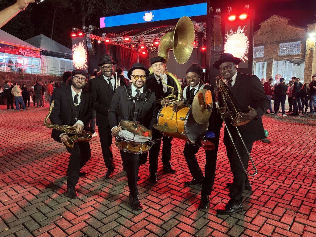 A imagem mostra uma banda de metais com seis integrantes posando em uma praça de tijolos à noite. Todos os músicos estão vestidos com ternos pretos, camisas brancas, gravatas e chapéus estilo fedora. Eles seguram seus instrumentos, que incluem saxofone, tambor, sousafone, bumbo com prato e trombone. Ao fundo, há um palco iluminado com luzes vermelhas e um banner que diz "FESTA DAS NAÇÕES", sugerindo que o evento é uma celebração cultural. Também é possível ver algumas pessoas, uma tenda branca e prédios de tijolos ao redor, criando um ambiente urbano e festivo. A iluminação vermelha dá um tom acolhedor e vibrante à cena, reforçando o clima de festa e música ao vivo.