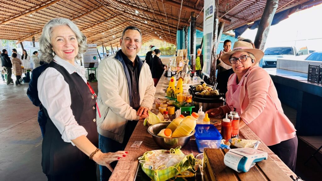 A imagem mostra três pessoas posando atrás de uma mesa em um ambiente coberto, possivelmente um mercado ou feira. * À esquerda, uma mulher de cabelos grisalhos veste uma camisa branca com colete preto. No centro, um homem sorridente usa uma jaqueta clara sobre uma camisa escura. À direita, uma mulher de chapéu, óculos e blazer rosa inclina-se sobre a mesa. * A mesa está repleta de produtos, incluindo garrafas com líquidos amarelados (provavelmente suco de milho), tigelas com pamonhas, condimentos e uma máquina de cartão de crédito. * O fundo revela uma estrutura de telhado de palha, outras pessoas circulando ao longe e um banner parcial com a palavra "PIRA" (referência a Piracicaba), indicando um evento público ou comunitário. A iluminação é natural e o clima parece ser de descontração e interação social.