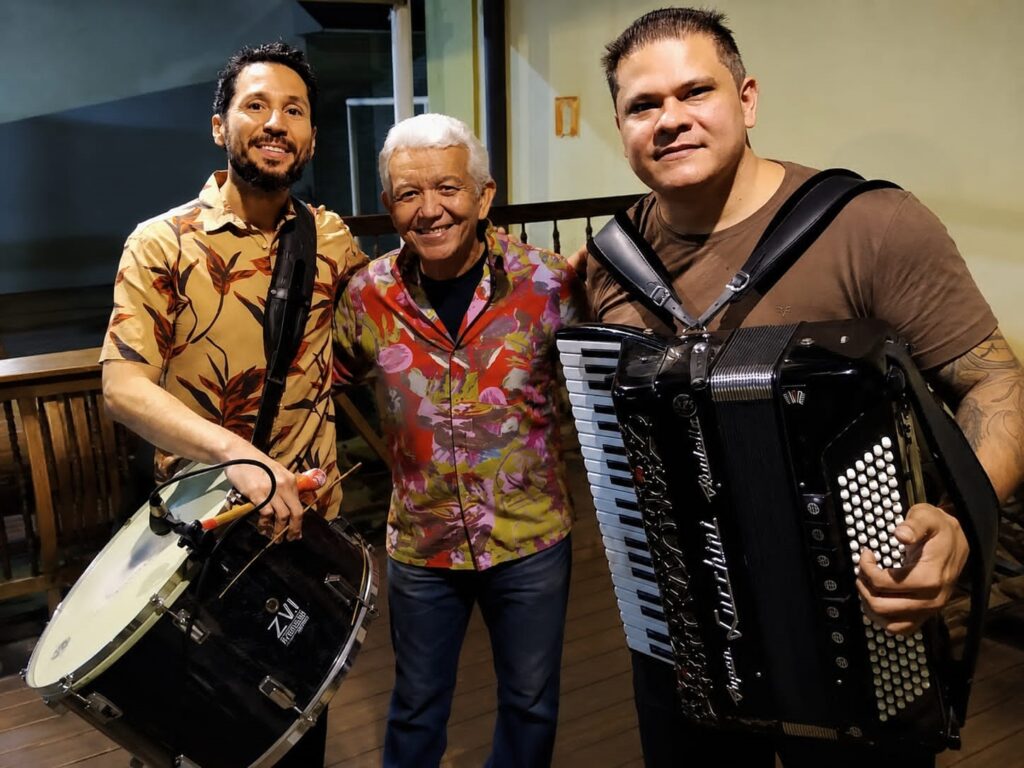 A imagem mostra três homens posando para uma foto juntos, provavelmente músicos. O homem à esquerda tem barba e veste uma camisa bege com estampa de folhas marrons, além de carregar um grande tambor preto com a marca "ZVI" visível. O homem ao centro é mais velho, com cabelos brancos, veste uma camisa colorida com estampa floral vibrante e calça jeans azul. O homem à direita tem uma tatuagem no braço esquerdo, usa uma camiseta marrom e segura um grande acordeão preto da marca "Lucchini". Eles estão em um ambiente com piso de madeira e uma estrutura de madeira ao fundo, com iluminação quente, possivelmente artificial. Os três sorriem para a câmera, e a foto foi tirada na altura da cintura, capturando-os do meio para cima.