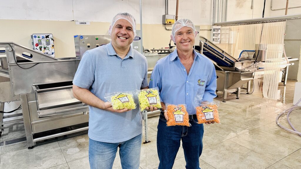Esta imagem mostra dois homens sorridentes em uma área de processamento de alimentos, orgulhosamente exibindo produtos embalados da cooperativa. As Pessoas: Ambos estão usando redes de proteção para cabelo (toucas descartáveis), indicando que estão em um ambiente de manipulação de alimentos. O homem à esquerda veste uma camisa polo azul claro e jeans; o da direita usa uma camisa de botão azul claro com o logotipo "COOPHORT" e jeans. O Produto: Eles seguram pacotes de vegetais processados (picados e prontos para consumo). O homem da esquerda segura dois pacotes com o que parecem ser vegetais amarelos/claros (talvez chuchu ou abobrinha), e o da direita segura dois pacotes de vegetais alaranjados (como cenouras). O Ambiente: Ao fundo, vê-se uma instalação industrial limpa, com piso claro, uma placa escrito "LAVAGEM" e equipamentos de aço inoxidável utilizados para processar ou higienizar os alimentos, reforçando o contexto profissional de produção hortifrúti.