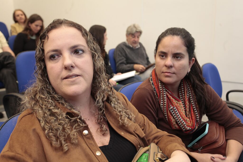 Nesta imagem, vemos um grupo de pessoas em um ambiente de reunião ou auditório. • Em primeiro plano: Duas mulheres estão sentadas lado a lado em cadeiras azuis. A da esquerda tem cabelos cacheados e veste uma jaqueta marrom. A da direita tem cabelos escuros presos e veste uma blusa marrom com um cachecol estampado colorido, segurando um celular. • Em segundo plano: Outras pessoas estão sentadas, também prestando atenção. É possível ver um homem de cabelos grisalhos e suéter cinza lendo ou observando algo, além de outras mulheres mais ao fundo. • Ambiente: O espaço parece ser uma sala de conferências ou curso, com iluminação interna e paredes de cor clara.