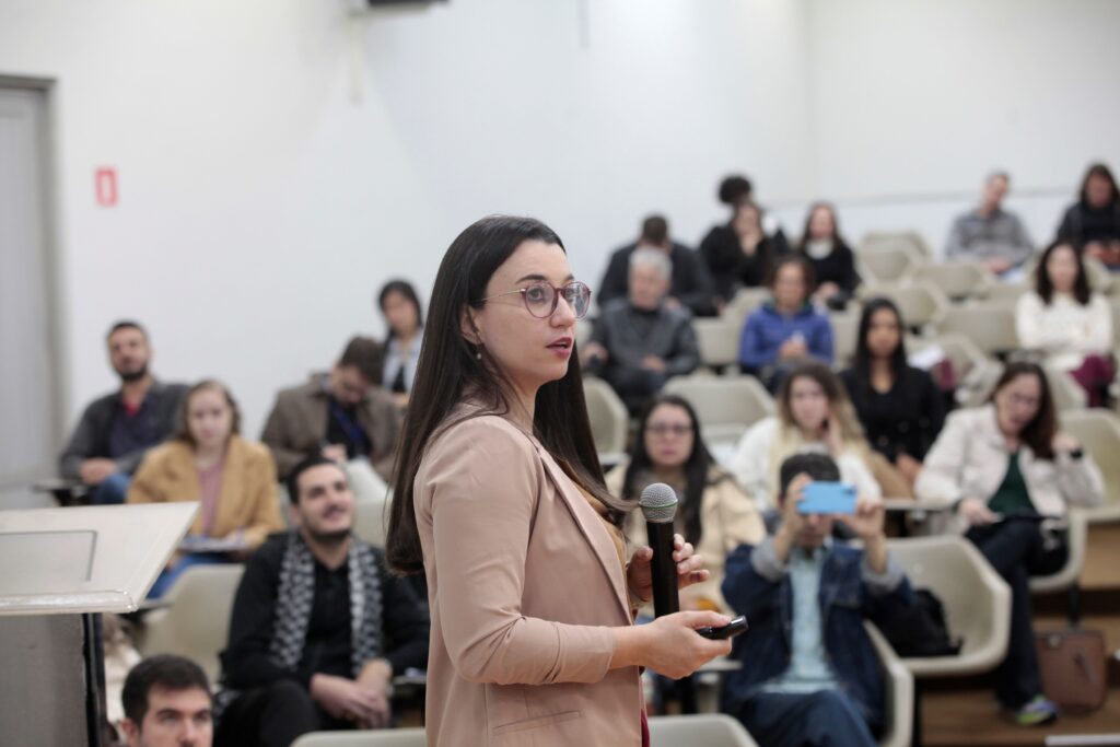 A imagem mostra uma pessoa fazendo uma apresentação ou palestra em um auditório ou sala de aula universitária. Descrição detalhada: Em primeiro plano, há uma mulher de cabelos longos e escuros, usando óculos e um blazer bege. Ela segura um microfone em uma das mãos e um pequeno controle remoto na outra, sugerindo que está apresentando slides ou conduzindo uma exposição. Sua expressão parece atenta e concentrada, como se estivesse explicando um tema ao público. Ao fundo, vê-se uma plateia sentada em fileiras de assentos escalonados, observando a apresentação. Algumas pessoas parecem estar acompanhando atentamente, enquanto uma pessoa na frente está usando um celular para fotografar ou gravar o momento. O ambiente é amplo, iluminado e com aparência acadêmica ou profissional, adequado para palestras, seminários, conferências ou aulas.