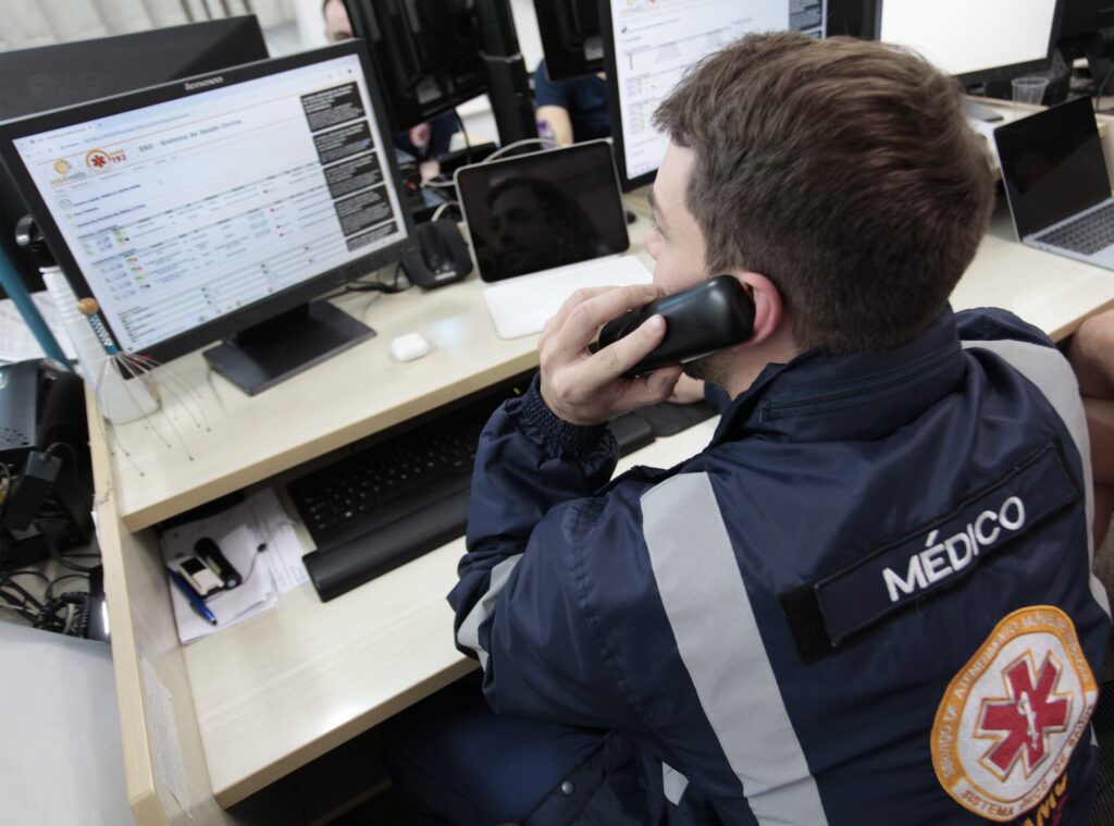A imagem mostra um profissional médico, identificado pela palavra "MÉDICO" nas costas do seu jaleco azul escuro, trabalhando em um ambiente de atendimento emergencial. Ele está sentado em frente a uma mesa, usando um telefone de linha fixa para se comunicar. Na mesa há vários equipamentos eletrônicos, incluindo um teclado, um tablet, um monitor de computador e um mouse. Na tela do computador, é possível ver um sistema online chamado "sys4web" do "SAMU 192", que é o Serviço de Atendimento Móvel de Urgência no Brasil. O ambiente parece ser uma central de atendimento ou despacho, onde o médico está provavelmente coordenando atendimentos de emergência. A iluminação é clara e o ambiente parece organizado, com foco na tecnologia para auxiliar no trabalho do profissional.