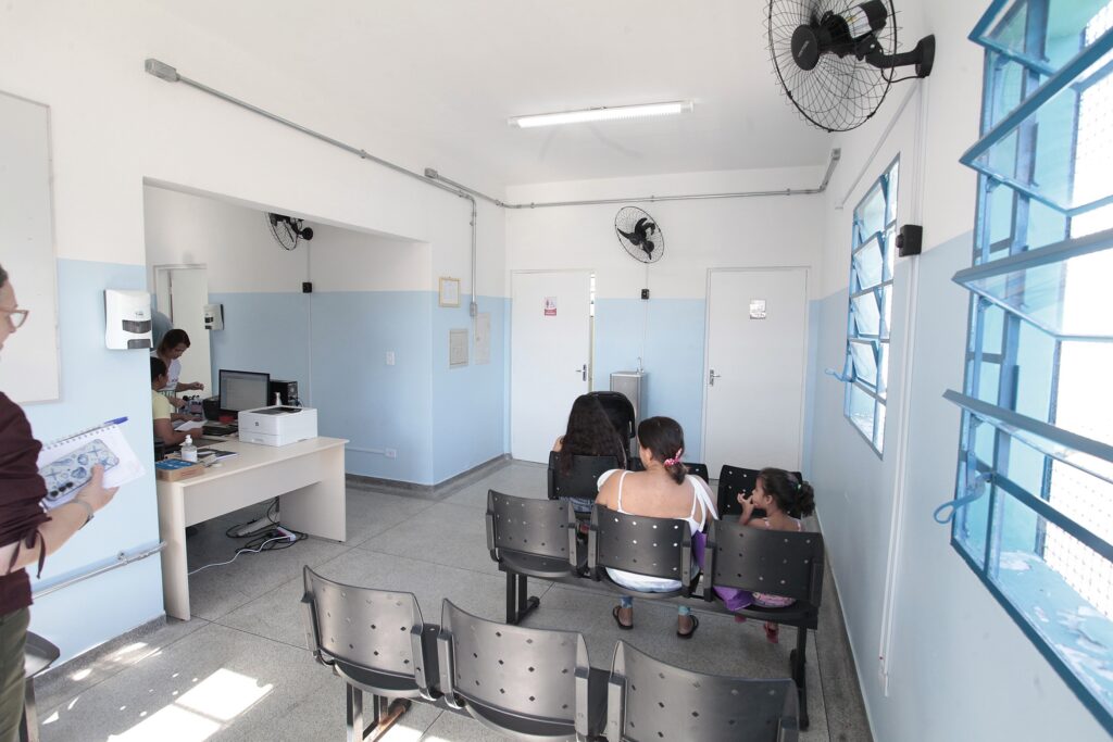 Uma sala de espera de um posto de atendimento ou consultório médico. Ambiente: O espaço é funcional e simples, com paredes brancas e uma faixa pintada de azul claro na parte inferior. O chão é de granilite cinza. Pessoas: No centro da sala, há três fileiras de cadeiras plásticas pretas. Uma mulher sentada, de costas, veste uma regata branca e cuida de uma criança ao seu lado. Atrás delas, outra pessoa está sentada. No canto esquerdo, há uma mesa de atendimento onde uma funcionária trabalha em um computador, com uma impressora ao lado. Detalhes: Há ventiladores de parede instalados no teto, portas brancas fechadas ao fundo e janelas de grade azul que permitem a entrada de luz natural. O ambiente transmite a sensação típica de uma unidade de saúde, organizada e iluminada.