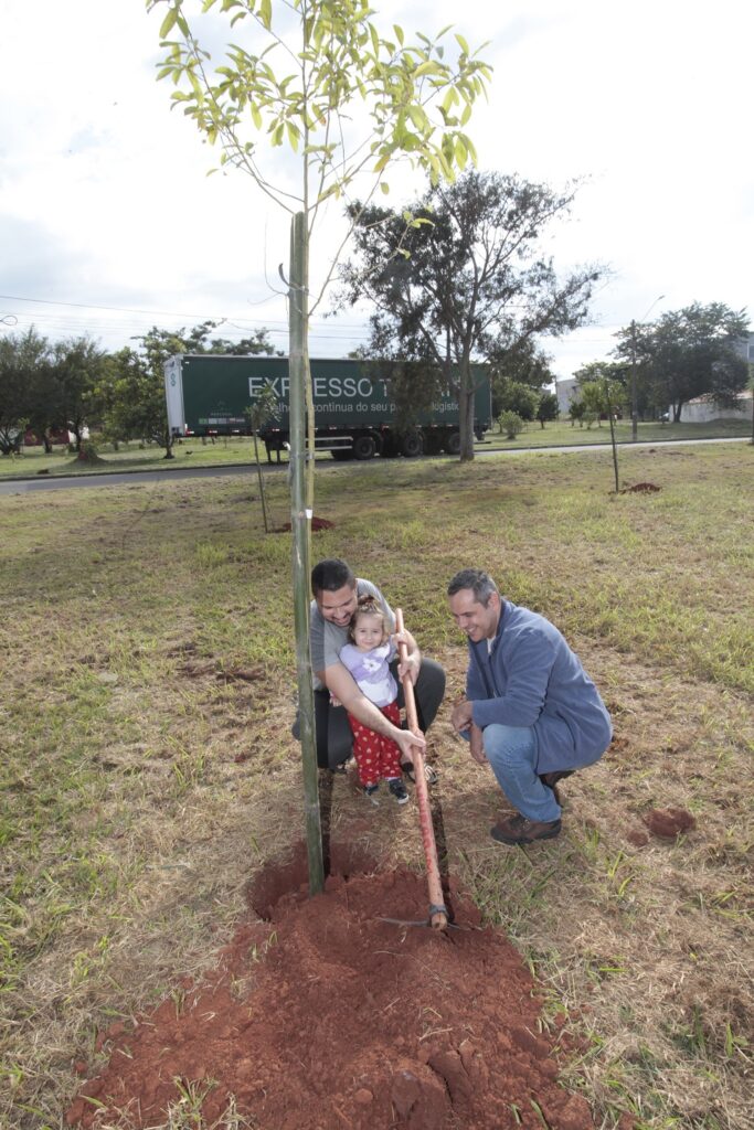 Nesta imagem, dois homens e uma criança pequena estão participando de uma ação de plantio de uma árvore em um terreno aberto e gramado. No centro da cena, agachados ao redor de uma muda de árvore recém-plantada (que está fixada a uma estaca de bambu): • Um homem de camiseta cinza está ajudando uma menina pequena, que veste uma blusa branca e calça estampada, a segurar um cabo de ferramenta de jardinagem para ajeitar a terra. • Ao lado deles, um homem vestindo um casaco azul, calça jeans e botas marrons observa a atividade, sorrindo. O solo em volta da muda está revolvido, mostrando a terra vermelha. Ao fundo, uma estrada separa o terreno de uma área arborizada, onde um caminhão verde de carga, com a inscrição "EXPRESSO T...", está passando. O céu está nublado, e o ambiente é uma área aberta e espaçosa.