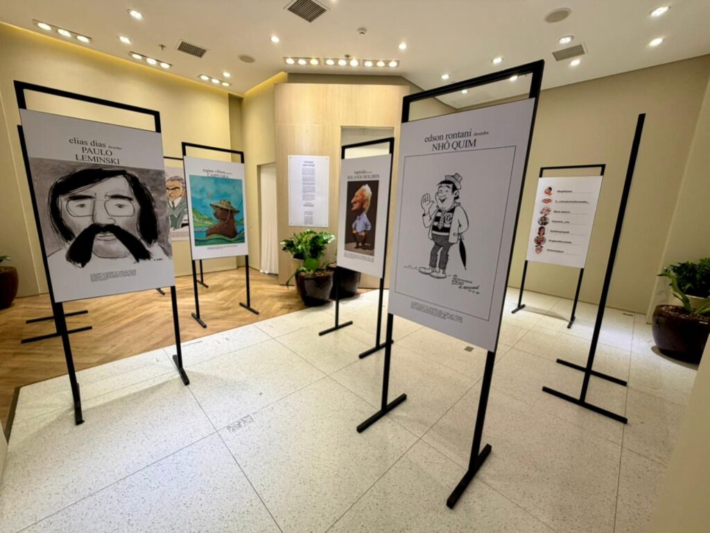 A foto mostra uma exposição de caricaturas e ilustrações em um ambiente interno bem iluminado, que parece um hall de shopping ou centro cultural.Detalhes da cena:Espaço: Salão amplo com piso de cerâmica clara e parte em piso de madeira. Teto com iluminação de spots embutidos. Vasos com plantas grandes decoram o local, deixando o clima mais acolhedor.Painéis expositivos: Vários totens pretos com banners brancos estão dispostos em zigue-zague pelo salão. Cada um traz a arte de um ilustrador e o personagem homenageado.Obras em destaque: Primeiro plano esquerdo: "elias dias desenha PAULO LEMINSKI" - retrato em preto e branco do poeta, com óculos, barba e bigode marcantes.Primeiro plano direito: "edson rontani desenha NHÔ QUIM" - caricatura colorida do personagem folclórico piracicabano, acenando e usando camisa do XV de Piracicaba.Ao fundo: Dá pra ver outros painéis, como "regina villares desenha CAPIVARA" com uma ilustração colorida, e "benedito... desenha ROLANDO BOLDIRIN" com um retrato de homem mais velho.Estilo: A mostra mistura traços realistas, caricatura e cores, homenageando figuras da cultura, literatura e identidade regional.É uma exposição organizada e curada, valorizando artistas e personagens locais. O layout convida o visitante a caminhar e conhecer cada painel.