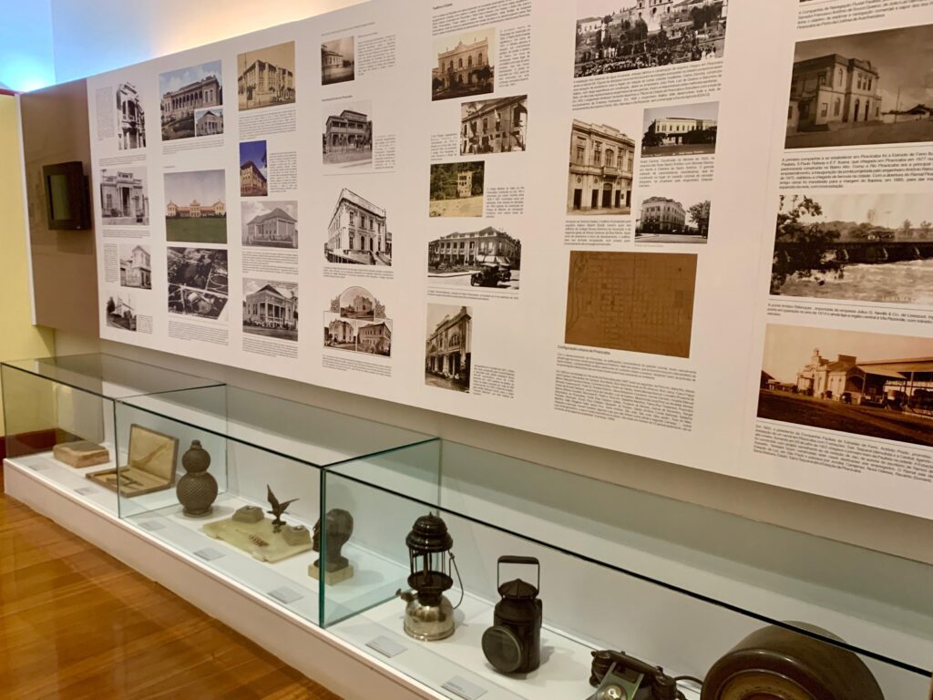 A imagem mostra uma exposição museológica dedicada à história urbana de Piracicaba, possivelmente localizada em um museu ou centro cultural. A parede principal é branca e repleta de fotografias históricas em preto e branco, apresentando cenas antigas da cidade, como teatros, clubes, escolas e infraestrutura urbana, além de textos explicativos em português que detalham a evolução da cidade, destacando acontecimentos e construções importantes entre os séculos XIX e XX. Na parte inferior da parede, há uma longa vitrine de vidro que exibe diversos artefatos históricos, incluindo lanternas antigas, objetos de metal e outros itens de época, que complementam o conteúdo visual e textual da parede. O ambiente possui um piso de madeira clara e iluminação focada, que valoriza os detalhes das imagens e dos objetos expostos. A composição transmite um ambiente educativo e informativo, com uma organização clara que permite ao visitante compreender a transformação urbana e cultural de Piracicaba ao longo do tempo, unindo documentos visuais, textos e objetos históricos em um único espaço expositivo.