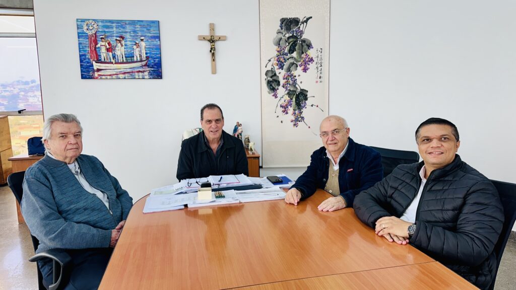 Quatro homens se reúnem em uma sala de escritório formal e bem iluminada, onde o ambiente de trabalho se mescla com elementos de arte e religiosidade.

A imagem mostra quatro homens de diferentes idades sentados ao redor de uma mesa retangular de madeira clara, em um ambiente que parece ser um escritório ou sala de reuniões. Do lado esquerdo da imagem, há um homem mais velho, com cabelos grisalhos e curtos, vestindo um suéter azul-cinza com zíper sobre uma camisa clara. Ele está sentado em uma cadeira de escritório escura e olha diretamente para a câmera com uma expressão séria, mas calma.

Ao lado dele, do outro lado da mesa, está um homem de meia-idade com cabelos escuros e encaracolados, vestindo uma jaqueta escura aberta sobre uma camisa. Ele sorri para a câmera. Na frente dele, sobre a mesa, há uma pilha de documentos, um porta-canetas com canetas e um pequeno objeto escuro que parece ser um grampeador ou suporte para celular. Atrás dele, em uma estante ou balcão baixo, pode-se ver uma pequena estatueta que representa a Virgem Maria com o Menino Jesus.

Continuando para a direita, há outro homem mais velho, calvo, com óculos, vestindo uma jaqueta escura sobre um suéter bege ou marrom claro. Ele olha para a câmera com um sorriso gentil. Suas mãos estão apoiadas sobre a mesa.

Por fim, no canto inferior direito, está um homem mais jovem, com cabelos escuros e curtos, vestindo uma jaqueta preta acolchoada sobre uma camisa branca. Ele também sorri para a câmera, com as mãos cruzadas sobre a mesa, exibindo um relógio no pulso esquerdo.

A parede branca ao fundo está decorada com três itens distintos. No lado esquerdo, acima do homem mais velho sentado, há um quadro colorido que representa figuras em um barco no mar, com uma bandeira e uma coroa de flores. Acima do homem de meia-idade com cabelo escuro, há um crucifixo de madeira com Cristo. Mais à direita, na parede, há uma pintura em pergaminho vertical com desenhos de uvas e folhas em tons de roxo e verde, com algumas inscrições em caligrafia oriental.

Perto do homem mais velho à esquerda, há uma grande janela que oferece uma vista parcialmente visível de uma área urbana com edifícios e vegetação sob um céu claro. A sala é bem iluminada, sugerindo luz natural vinda da janela e talvez iluminação artificial não visível na imagem.

Não há animais na imagem. A atmosfera geral é de formalidade e cordialidade. As expressões sorridentes da maioria dos participantes sugerem um encontro amigável ou uma reunião de trabalho bem-sucedida, com um tom respeitoso e profissional. O cenário denota um ambiente de seriedade e cultura, com toques pessoais através das obras de arte e objetos religiosos.