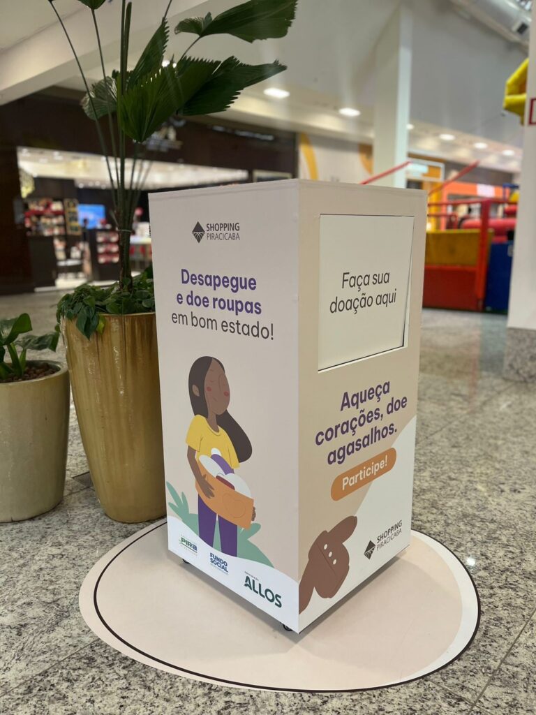 A imagem mostra uma caixa de coleta para doação de roupas localizada dentro de um shopping, especificamente no Shopping Piracicaba. A caixa é branca, retangular e possui um espaço para depositar as doações no topo lateral. Na frente, há uma ilustração de uma mulher segurando uma caixa com roupas e o texto em português que incentiva as pessoas a doarem roupas em bom estado: "Desapegue e doe roupas em bom estado!" e "Aqueça corações, doe agasalhos. Participe!". A caixa também exibe o nome do shopping, "Shopping Piracicaba", e os logos de entidades envolvidas, como o Fundo Social e a administração Allos. Ao lado da caixa, há um vaso grande com uma planta, e o ambiente ao redor indica que é uma área interna do shopping, com piso de pedra polida e áreas comerciais ao fundo, além de um espaço infantil visível. A iluminação é clara e típica de um espaço comercial fechado. A caixa está posicionada de forma a chamar a atenção dos frequentadores para incentivar a doação de roupas.