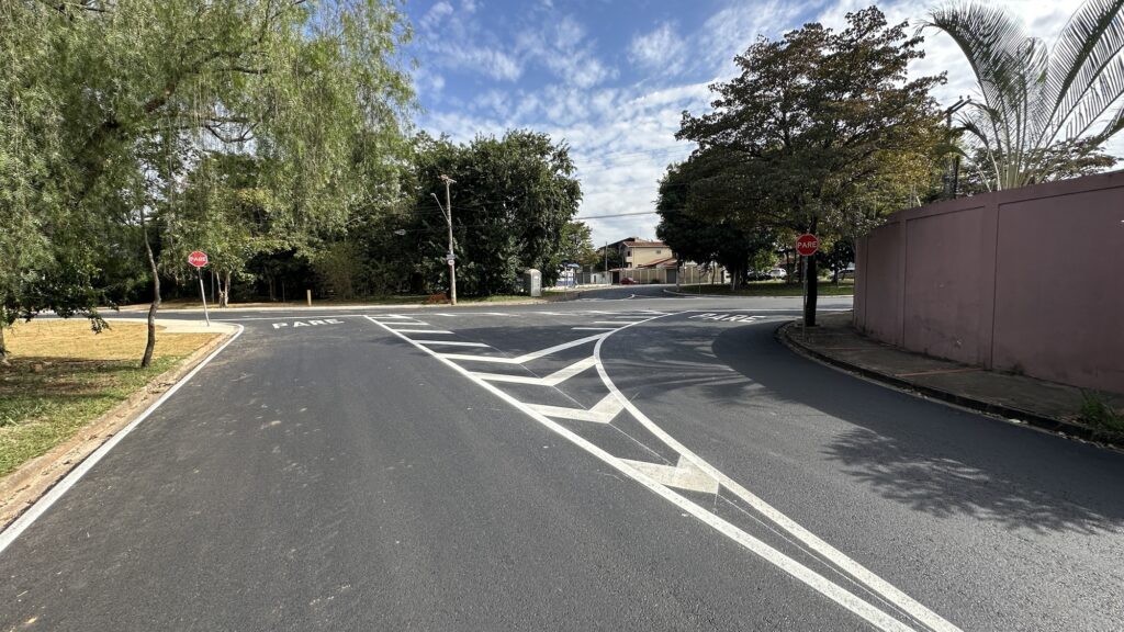 A imagem mostra um cruzamento em forma de T em uma estrada asfaltada, com sinalização clara no chão e placas de "PARE" em ambos os lados da via que se encontra perpendicular à estrada principal. O céu está claro com algumas nuvens e a luz do sol ilumina a cena, criando sombras suaves. Há árvores verdes ao redor e um muro de cor clara à direita da imagem. A estrada está vazia, sem veículos ou pessoas, transmitindo uma sensação de tranquilidade e ordem. A sinalização indica que os veículos que chegam pela via lateral devem parar antes de entrar na estrada principal.