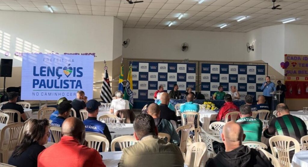 A imagem mostra um evento realizado em um salão amplo e bem iluminado, possivelmente uma conferência, reunião ou cerimônia oficial. Na parte frontal do ambiente, há um palco com um painel ao fundo exibindo repetidamente o logo da Prefeitura de Lençóis Paulista. À direita do palco, uma pessoa está falando em um púlpito, provavelmente fazendo uma apresentação ou discurso. O público está sentado em mesas redondas, e muitos participantes vestem camisetas com nomes de cidades, como "Pratânia" e "Macatuba", indicando que são representantes ou grupos dessas localidades. Há também duas bandeiras posicionadas próximas ao palco, uma delas sendo a bandeira do Brasil. Na parede à esquerda, um telão grande exibe o brasão da Prefeitura de Lençóis Paulista. O ambiente transmite uma atmosfera formal, porém acolhedora, sugerindo um encontro comunitário ou evento governamental voltado para a população local. No painel do fundo, há frases motivacionais como "O cuidado não envelhece" e "Cada idade tem sua beleza", indicando que o evento pode estar relacionado com temas de cuidado, saúde ou bem-estar, possivelmente focado em diferentes faixas etárias.