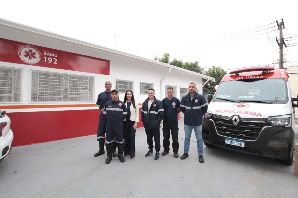 A imagem mostra um grupo de seis pessoas posando em frente a uma base do SAMU (Serviço de Atendimento Móvel de Urgência). * No centro, há uma mulher vestindo um blazer claro e calça escura, cercada por cinco homens. * Quatro dos homens vestem uniformes profissionais azul-marinho com faixas refletivas, característicos das equipes do SAMU, enquanto um homem no meio veste roupas casuais, incluindo uma jaqueta escura. * À direita, vê-se uma ambulância Renault branca e vermelha, com a palavra "AMBULÂNCIA" escrita de forma invertida no capô e a placa TJW7J80. * Ao fundo, há um edifício com paredes brancas e uma faixa horizontal vermelha, onde se lê o logotipo do SAMU e o número "192". O ambiente parece ser um pátio externo pavimentado.