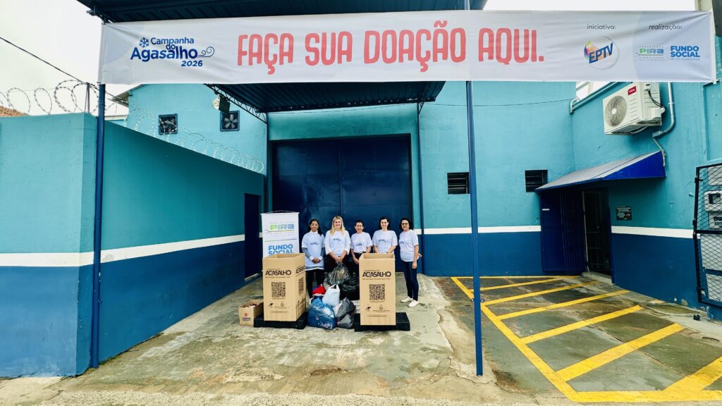 A imagem mostra uma campanha de arrecadação de agasalhos intitulada "Campanha do Agasalho 2026". Cinco mulheres estão posando em frente a um prédio pintado em tons de azul, todas vestindo camisetas brancas com a identidade visual da campanha. Elas estão atrás de duas grandes caixas de papelão, que estão cheias de roupas doadas, acondicionadas em sacos plásticos. Acima delas, há uma faixa branca com a frase "FAÇA SUA DOAÇÃO AQUI." em letras grandes, indicando o local para entrega das doações. No banner também estão os logos e nomes dos organizadores da campanha: EPTV, Prefeitura de Piracicaba e Fundo Social de Solidariedade. O ambiente é externo, com piso de concreto e uma parede azul ao fundo. A campanha incentiva a doação com a frase "Doe com amor. Faz bem fazer o bem." A imagem transmite uma sensação de solidariedade e engajamento comunitário para ajudar pessoas que precisam de agasalhos.