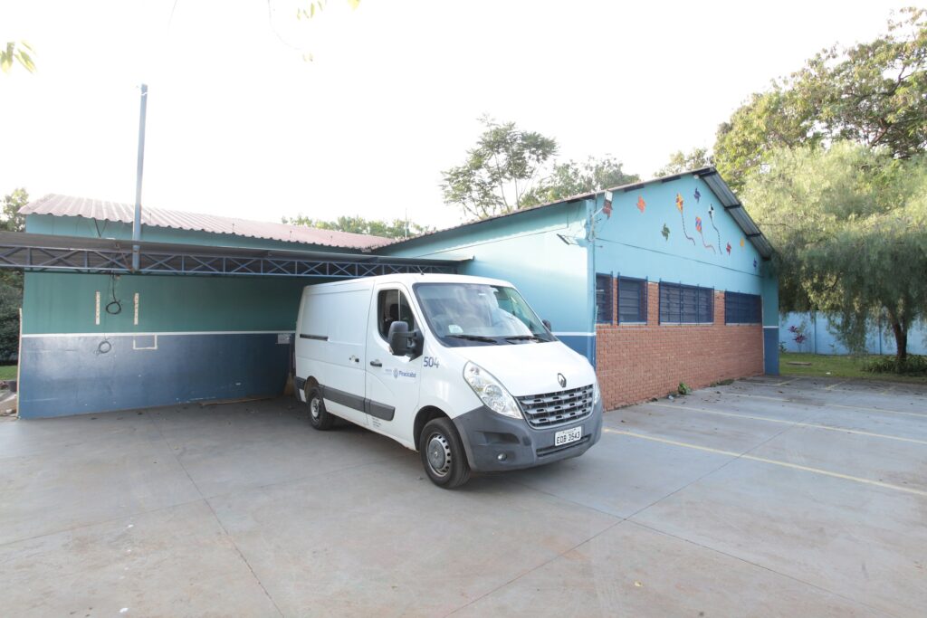 Na imagem, vemos uma van branca da Renault estacionada diante de um prédio azul e verde com base em tijolos vermelhos. A construção possui telhado de metal ondulado e janelas com molduras azuis, além de pinturas coloridas de pipas na parte superior, que dão um aspecto alegre e comunitário ao espaço. A van traz inscrições como o número “504” e o nome “Piracicaba”, além da placa “EOB 3543”. O cenário é um estacionamento pavimentado cercado por árvores e vegetação, transmitindo a atmosfera de utilidade urbana em contraste com o caráter lúdico da arquitetura comunitária.
