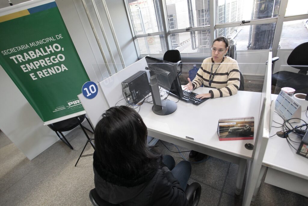 A imagem mostra um ambiente de atendimento ao público, possivelmente um escritório da Secretaria Municipal de Trabalho, Emprego e Renda. * No centro, uma atendente de óculos e blusa listrada está sentada atrás de uma mesa branca, digitando em um computador e gesticulando enquanto fala com uma pessoa sentada à sua frente. * Entre elas, há uma divisória de acrílico transparente para proteção. * À esquerda, um banner verde identifica o órgão público e há uma placa circular azul com o número "10" indicando o posto de atendimento. * Sobre a mesa, além do computador, observam-se um calendário de mesa com a foto de um carro e um calendário de parede (ou suporte) com o ano de 2026. * O ambiente possui grandes janelas com persianas ao fundo, permitindo a entrada de luz natural, e o piso é de granilite. O foco da cena é a interação profissional entre a atendente e o cidadão.