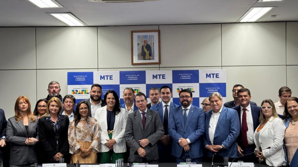 A imagem retrata um grupo maior de pessoas, composto por homens e mulheres, posando para uma foto em um auditório ou sala de reuniões. Eles estão posicionados em frente a um painel com as logos do "Ministério do Trabalho e Emprego" e "Governo Federal", e há um retrato oficial do presidente do Brasil na parede. O ambiente parece ser uma reunião ou evento institucional oficial.