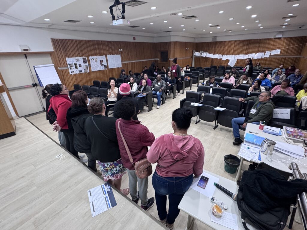 A imagem mostra uma reunião ou evento em uma sala ampla e moderna, provavelmente uma conferência ou workshop. No primeiro plano, há várias pessoas em pé, vistas de costas, algumas conversando entre si. Entre elas, destaca-se uma mulher com um moletom rosa e outra com uma saia floral. À frente, há várias pessoas sentadas em cadeiras pretas ergonômicas, dispostas em fileiras, algumas prestando atenção, outras conversando. A sala tem paredes de madeira clara e o chão é claro também, com iluminação forte e uniforme proveniente do teto, onde está instalado um projetor. Sobre uma das cadeiras, há uma placa com a palavra "QUEBRADA" e, abaixo, "ORGANIZAÇÃO FINANCEIRA", sugerindo que o tema do evento está relacionado a finanças. O ambiente é informal, com pessoas vestidas casualmente, indicando um encontro comunitário, educacional ou de treinamento. A disposição dos móveis e o número de participantes mostram que é um espaço preparado para interação e aprendizado em grupo.