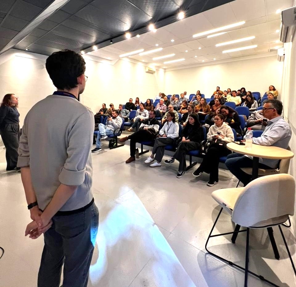 Nesta imagem, vemos um ambiente de auditório ou sala de conferências durante uma apresentação ou palestra. • Primeiro plano: Uma pessoa de costas, vestindo uma blusa de lã bege e calça escura, está de pé, observando a cena. Ao lado dela, à esquerda, uma mulher em pé também observa o grupo. • O público: Há um grupo de pessoas sentadas em fileiras de cadeiras azuis, prestando atenção à apresentação. • Destaque: À direita, sentado à mesa, um homem de óculos e camisa clara parece ser o palestrante ou moderador. • Ambiente: A sala possui iluminação clara, teto com placas acústicas e é um espaço funcional, ideal para reuniões ou cursos.
