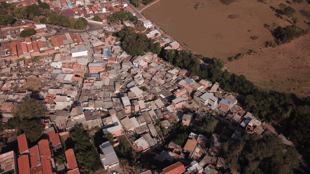 A imagem mostra uma vista aérea de um bairro residencial denso e semi-urbano, localizado ao lado de um campo aberto e seco. À esquerda, é possível ver muitas casas próximas umas das outras, com telhados em tons de terracota e cinza, construídas principalmente com tijolos vermelhos. As casas parecem estar em diferentes estágios de construção ou manutenção. À direita, há uma grande área de vegetação seca, com gramíneas amareladas, indicando um ambiente árido ou em estação seca. A luz do sol está baixa, sugerindo o final da tarde, e cria sombras longas que dão um tom quente à cena. Uma rua estreita e pavimentada passa pela parte superior esquerda da imagem, e não há pessoas visíveis, conferindo um aspecto tranquilo e silencioso ao local. A imagem destaca o contraste entre a área urbana densa e o campo aberto ao lado.