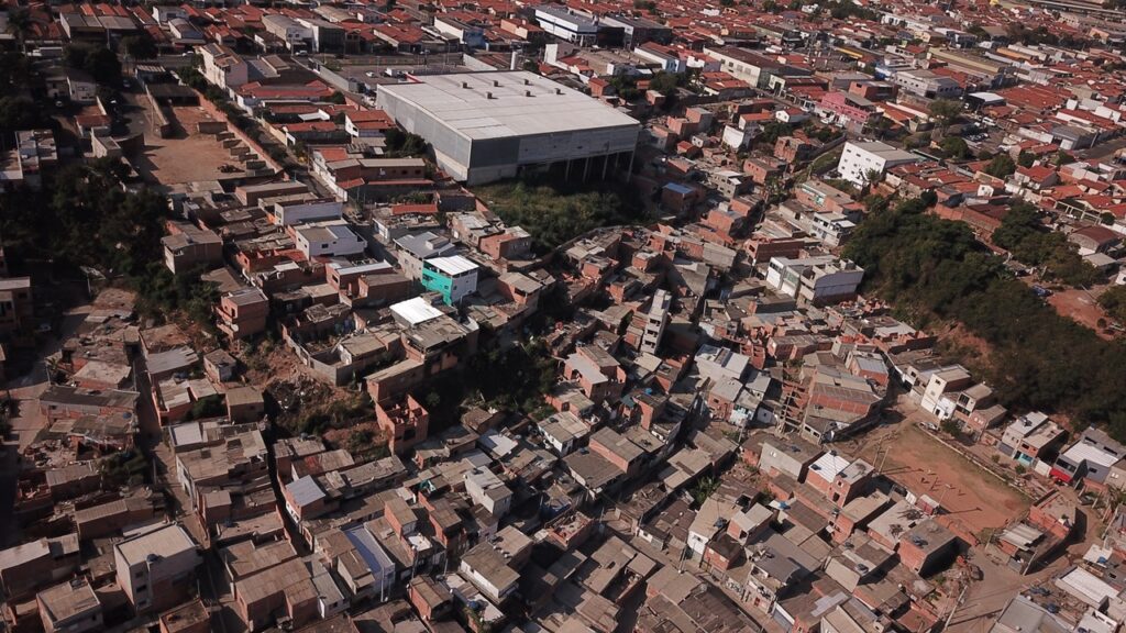 A imagem mostra uma vista aérea de uma área urbana densamente povoada, provavelmente uma favela ou comunidade informal em Piracicaba. No centro superior da imagem, há um grande edifício moderno, possivelmente industrial ou comercial, com estrutura mais organizada e telhados claros. Ao redor dele, há uma grande concentração de casas pequenas, construídas de forma irregular e muito próximas umas das outras, com telhados variados em tons de cinza, marrom e terracota. As ruas são estreitas e sinuosas, e a infraestrutura parece desordenada. Pouca vegetação é visível, com apenas algumas pequenas áreas verdes dispersas. A iluminação forte indica que a foto foi tirada durante o dia, com o sol alto, criando sombras marcadas. A imagem evidencia o contraste entre a construção planejada do edifício maior e a ocupação densa e improvisada das moradias ao redor.