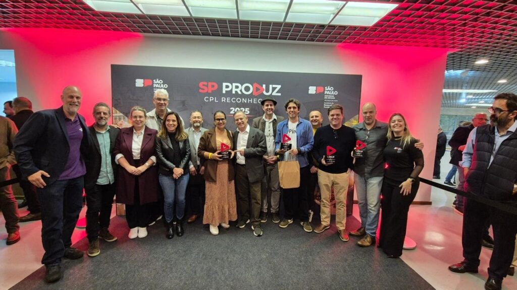 Esta foto registra um evento de premiação chamado "SP PRODUZ - CPL RECONHECE 2025". O Grupo: Vemos um grupo grande de pessoas, formado por homens e mulheres de idades variadas, posando para uma foto oficial em frente a um painel (backdrop) do evento. A Premiação: Algumas pessoas no centro e à direita do grupo seguram troféus personalizados com um formato triangular vermelho, indicando que são os premiados ou representantes das iniciativas reconhecidas. O Ambiente: É um evento formal, mas com clima descontraído. O local tem iluminação com tons avermelhados nas laterais, teto de grade metálica e piso claro. O painel ao fundo traz a marca do Governo do Estado de São Paulo e o título do evento em destaque. Vestimenta: A maioria das pessoas veste roupas estilo "business casual" ou casuais elegantes (camisas, blazers, jeans, calças de sarja), refletindo um ambiente corporativo ou institucional.