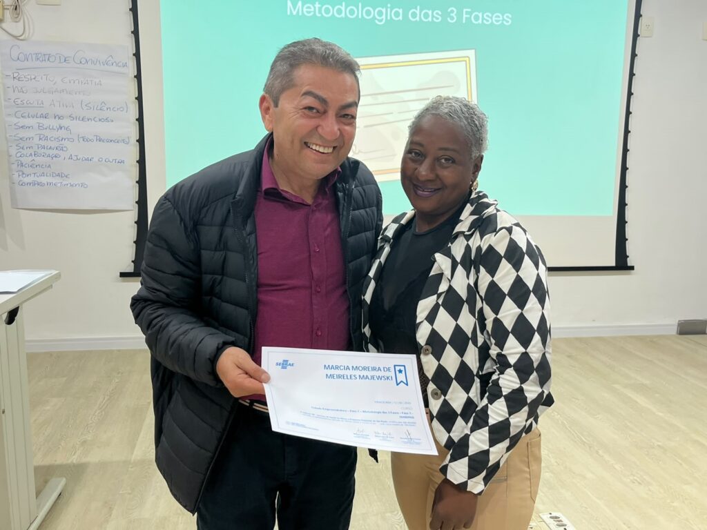 A imagem mostra duas pessoas, um homem e uma mulher, posando para a foto em um ambiente interno, possivelmente uma sala de aula ou treinamento. O homem está à esquerda, veste uma camisa vinho sob uma jaqueta preta acolchoada e sorri para a câmera. A mulher, à direita, usa um blazer xadrez preto e branco sobre uma blusa preta e calça bege, e segura um certificado do SEBRAE, que está parcialmente visível e indica que ela se chama Márcia Moreira de Meireles Majewski. Atrás deles, há um projetor exibindo o texto "Metodologia das 3 Fases" e, na parede, um cartaz com um "Contrato de Convivência" que lista regras como respeito, empatia, silêncio, sem bullying, colaboração, entre outras. O ambiente é iluminado e parece ser uma sala destinada a capacitações ou palestras.