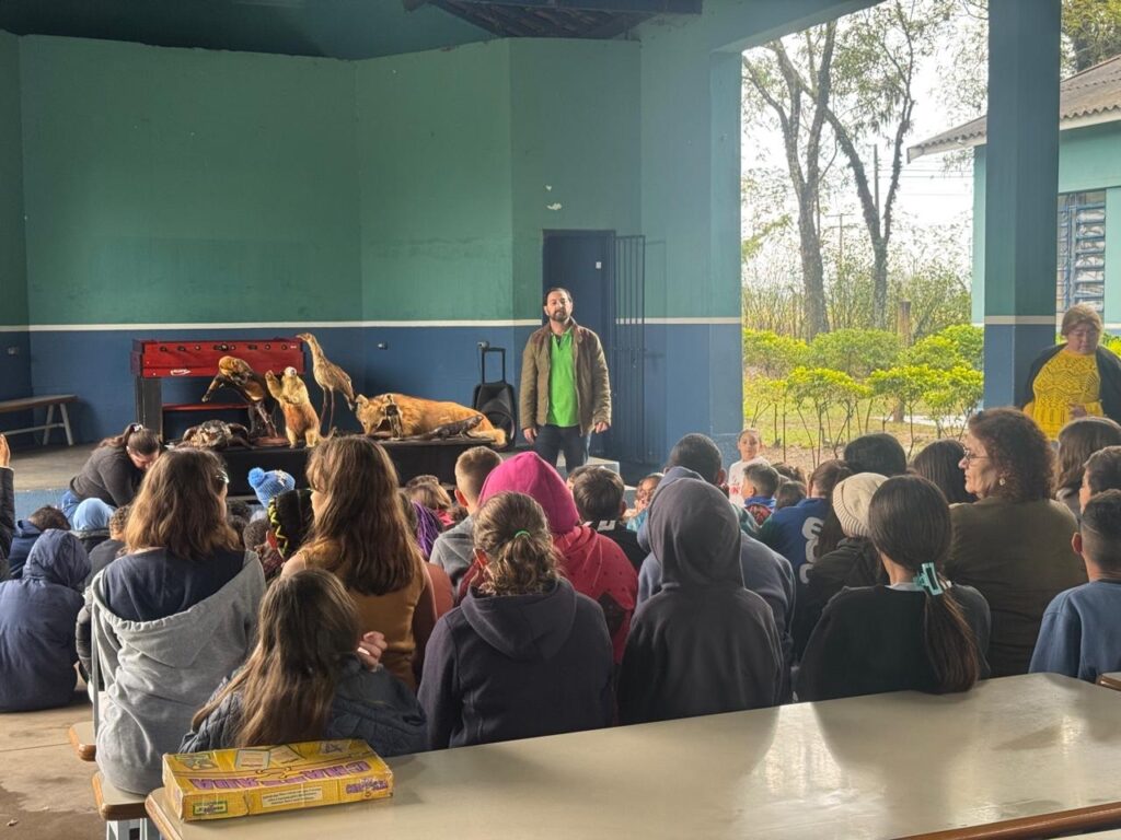 A imagem mostra uma apresentação educativa em um pavilhão aberto, com paredes pintadas de azul claro e uma faixa azul escuro. Um homem de barba, vestido com camiseta verde e jaqueta marrom, está em pé falando para um grupo de crianças e adultos sentados em bancos. Na mesa à sua frente, há vários animais empalhados, incluindo um pequeno macaco, um pássaro e um animal maior, possivelmente uma raposa ou um felino selvagem. Ao fundo, pode-se ver árvores e o ambiente externo. Na frente da imagem, há uma caixa de jogo de tabuleiro com o nome "CHALLENGE". O público está atento à apresentação, que parece ser sobre animais ou natureza. A iluminação é natural, típica de um dia nublado.