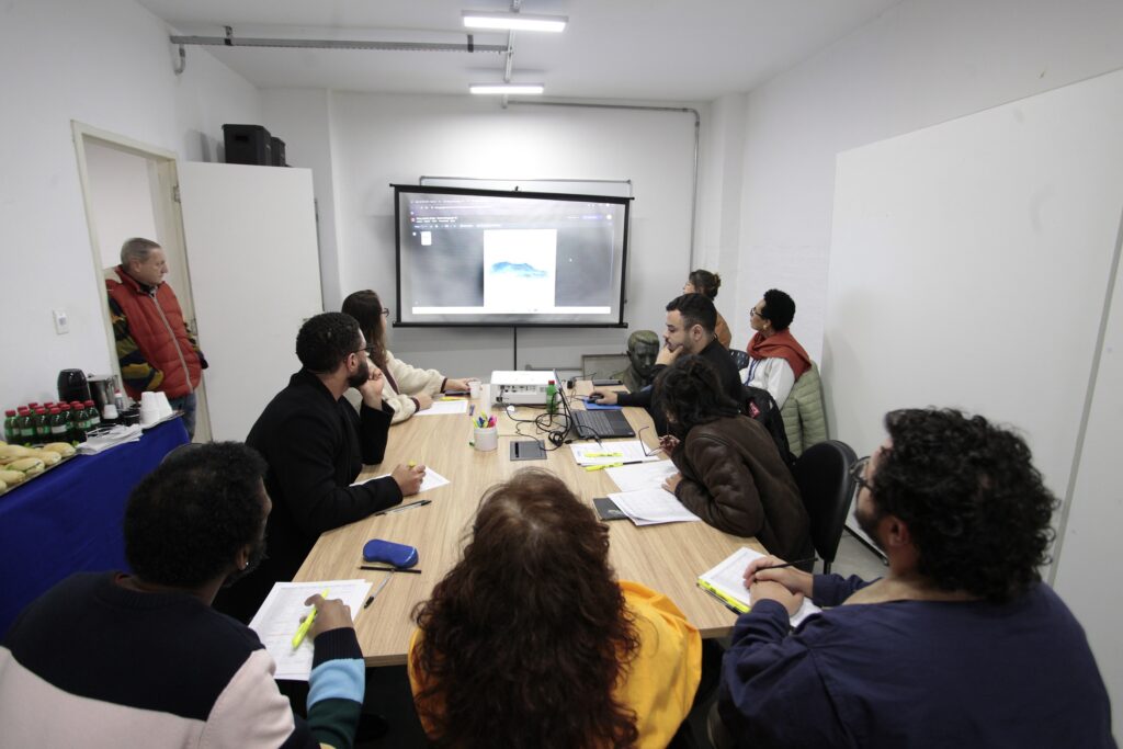 Na imagem, vemos um grupo de pessoas reunidas em torno de uma mesa retangular de madeira, em um ambiente de reunião ou sala de aula. Todos estão atentos a uma tela de projeção que exibe uma paisagem de montanha, enquanto sobre a mesa há cadernos, canetas, marcadores e um laptop conectado ao projetor. Ao lado, uma mesa coberta por toalha azul traz garrafas de refrigerante, copos e pães, sugerindo um apoio para os participantes. Um observador está de pé próximo à porta, acompanhando a sessão. A cena transmite a atmosfera de aprendizado colaborativo e discussão em grupo, destacando o engajamento coletivo em torno de conteúdos visuais.