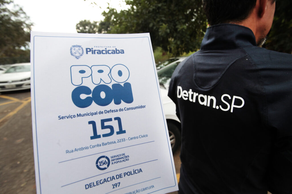 A imagem é um close com foco em um folheto informativo da Prefeitura de Piracicaba, com um agente do Detran.SP de costas ao fundo.Detalhes da cena:Primeiro plano - Folheto: Topo: Brasão e o texto "PREFEITURA DE Piracicaba".Destaque: Logo grande "PROCON" em azul e branco, com o subtítulo "Serviço Municipal de Defesa do Consumidor".Contatos: Número "151" em destaque, endereço "Rua Antônio Corrêa Barbosa, 2233 - Centro Cívico".Abaixo: Ícone do "156 SERVIÇO DE INFORMAÇÕES À POPULAÇÃO" e "DELEGACIA DE POLÍCIA 197". No rodapé aparece "DISTRIBUIÇÃO GRATUITA".Fundo: A parte de trás de um homem usando jaqueta preta com "Detran.SP" em letras brancas grandes. Ao fundo, desfocado, dá pra ver um estacionamento com carros e árvores.A foto mostra uma ação de divulgação dos canais de atendimento ao cidadão em Piracicaba, com parceria entre o Procon Municipal e o Detran.SP. O material é informativo e de acesso público.
