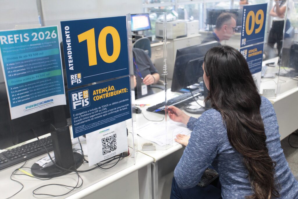 A cena: Uma atendente, vista de costas com longos cabelos escuros, está sentada em seu posto (Atendimento 10) trabalhando com documentos. À sua frente, há um painel de acrílico transparente para proteção. Informações em destaque: Há diversos avisos colados nas divisórias de vidro, detalhando as condições do REFIS 2026 para os contribuintes: Descontos: O cartaz à esquerda explica os descontos em juros e mora para dívidas de até R 500 m i l e a c i m a d e R 500mileacimadeR 500 mil, variando conforme o número de parcelas (de 2 a até 120 parcelas). Orientações: O cartaz central reforça a "Atenção, Contribuinte", informando que é obrigatória a inclusão de todos os débitos pendentes com a prefeitura, com exceção de pagamentos à vista. Conectividade: Há também um código QR para conexão Wi-Fi. Ambiente: O local parece ser um escritório administrativo bem estruturado, com computadores e outros atendentes visíveis ao fundo, reforçando a natureza de um serviço público de atendimento ao cidadão.