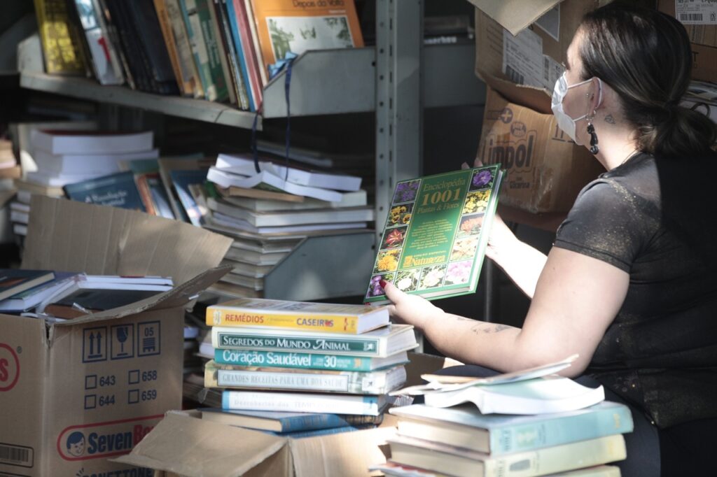 A imagem mostra uma mulher usando máscara, de perfil, organizando livros em um ambiente que parece ser um depósito ou uma biblioteca. Ela está segurando um livro verde com o título "Enciclopédia 1001 Plantas & Flores". Ao redor dela, há várias prateleiras metálicas e caixas de papelão cheias de livros. Uma das caixas tem a marca "Seven Boys" estampada. A iluminação é natural, entrando pela lateral, destacando os detalhes dos livros e da mulher. A cena transmite uma atmosfera de trabalho manual, organização e cuidado com os livros em um espaço de armazenamento.