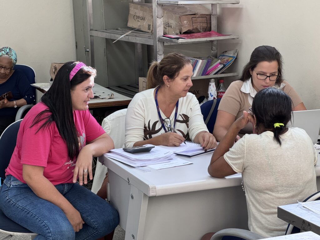 A imagem mostra um ambiente interno, possivelmente um escritório ou local de atendimento, onde quatro mulheres estão sentadas ao redor de uma mesa branca. Uma delas, vestindo uma camiseta rosa e calça jeans azul, está sentada à esquerda, olhando para o centro da mesa. Outra mulher, com uma blusa clara de manga longa com estampa escura, está escrevendo em um caderno, com uma calculadora e papéis sobre a mesa. Uma terceira mulher, usando óculos e uma blusa bege, observa a interação. A quarta mulher, com o cabelo preso e vestindo uma blusa clara, está com as costas para a câmera, participando da conversa. Ao fundo, uma senhora com lenço na cabeça é parcialmente visível. No ambiente, há uma estante metálica cheia de caixas, livros e uma garrafa de Coca-Cola. A iluminação é forte, provavelmente fluorescente, e o clima parece ser de trabalho ou atendimento administrativo.
