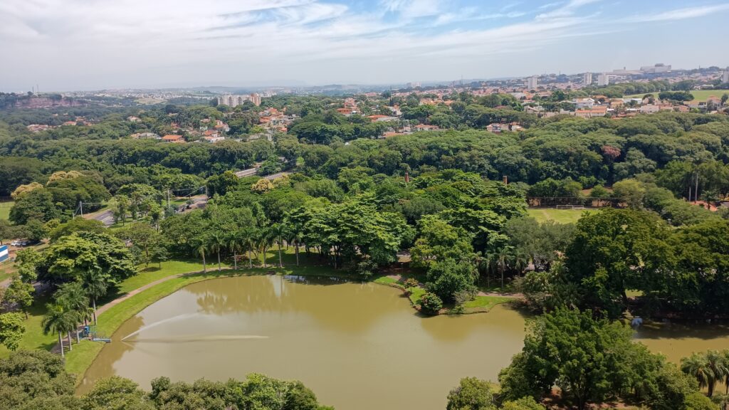 Esta imagem aérea captura uma vista panorâmica de um parque urbano em um dia ensolarado. * O elemento central é um lago de águas esverdeadas, que apresenta um pequeno chafariz lançando água na superfície. * Ao redor do lago, há uma densa área de vegetação com muitas árvores e palmeiras, criando um ambiente arborizado que contrasta com a área urbana ao fundo. * No horizonte, observa-se uma cidade com diversos edifícios, casas e ruas, indicando que o parque está localizado em uma zona residencial ou central densamente povoada. * O céu apresenta algumas nuvens finas e alongadas, sugerindo um clima agradável e aberto. A perspectiva elevada permite visualizar a transição entre a área de lazer natural e a infraestrutura da cidade.