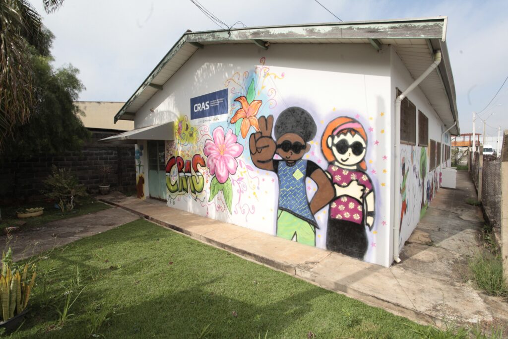A imagem mostra a lateral de um prédio que abriga o CRAS (Centro de Referência de Assistência Social) da cidade de Piracicaba. A parede do prédio está decorada com um mural colorido e vibrante, que ocupa praticamente toda a sua extensão. O mural apresenta figuras estilizadas e caricatas de duas pessoas: um homem com cabelo afro fazendo o sinal de paz com a mão e uma mulher de cabelo laranja, ambos usando óculos escuros. O fundo do mural é composto por formas geométricas e elementos florais em cores fortes como rosa, laranja, verde e azul. Acima do mural, há uma placa com o logo da Prefeitura de Piracicaba e o nome completo do CRAS. O prédio é simples, de cor clara, e está situado em uma área com gramado e uma calçada de concreto. Ao lado, é possível ver uma cerca de arame. A iluminação natural indica que a foto foi tirada durante o dia. O conjunto transmite uma sensação de acolhimento e alegria, refletindo a função social do CRAS.