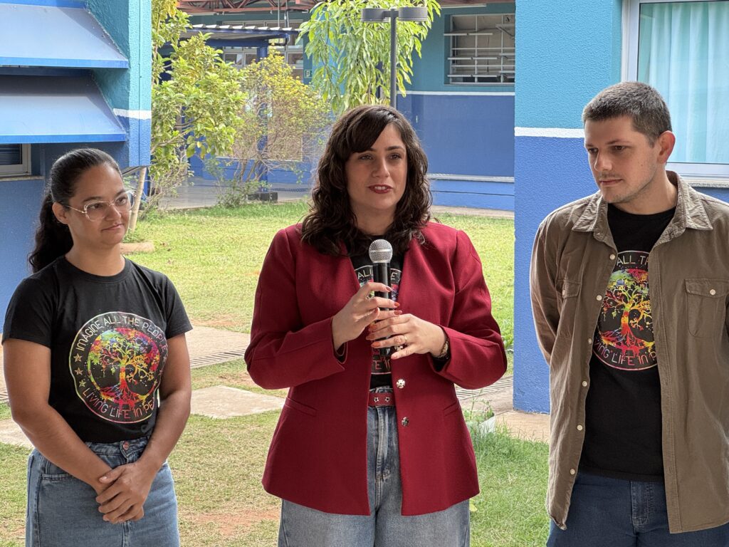 A imagem mostra três pessoas em um ambiente externo, possivelmente em um pátio ou área de escola. No centro, uma mulher está falando ao microfone, vestindo um blazer vermelho por cima de uma camiseta preta com uma estampa colorida de uma árvore e a frase "IMAGINE ALL THE PEOPLE LIVING LIFE IN PEACE" (Imagine todas as pessoas vivendo a vida em paz). Ao lado dela, à esquerda, está uma jovem mulher com cabelos longos e óculos, usando a mesma camiseta estampada, e à direita, um jovem homem com cabelo curto, também vestido com a mesma camiseta por baixo de uma camisa bege aberta. Ao fundo, vê-se uma construção azul com janelas e grades, além de árvores e gramado. A cena sugere que estão participando de um evento, talvez uma apresentação ou discurso relacionado à paz e convivência.