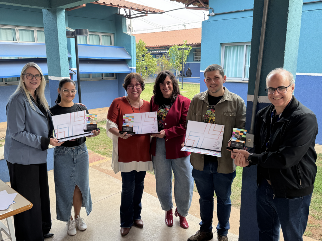 A imagem mostra um grupo de seis pessoas posando para uma foto em um ambiente externo, provavelmente em um campus ou área educacional, já que ao fundo há um gramado e estruturas que parecem prédios escolares. Todos estão alinhados lado a lado, segurando pastas brancas com o logo da "Faculdade Sebrae" e troféus pequenos, sugerindo que participaram de um evento ou receberam algum tipo de reconhecimento.
As pessoas vestem roupas casuais e semi-formais, com algumas usando blazers e outras camisetas. O cenário é composto por paredes pintadas de azul vibrante e um piso de concreto, com uma cobertura transparente acima, que deixa a luz natural iluminar o local de forma suave. No fundo, é possível ver uma área aberta com grama e mais construções.
O texto visível na pasta menciona "Faculdade Sebrae" e "Inovação e Sustentabilidade na Educação", indicando que o evento ou premiação está relacionado a esses temas. A atmosfera geral da imagem é de celebração e profissionalismo em um contexto educacional voltado para inovação e sustentabilidade.