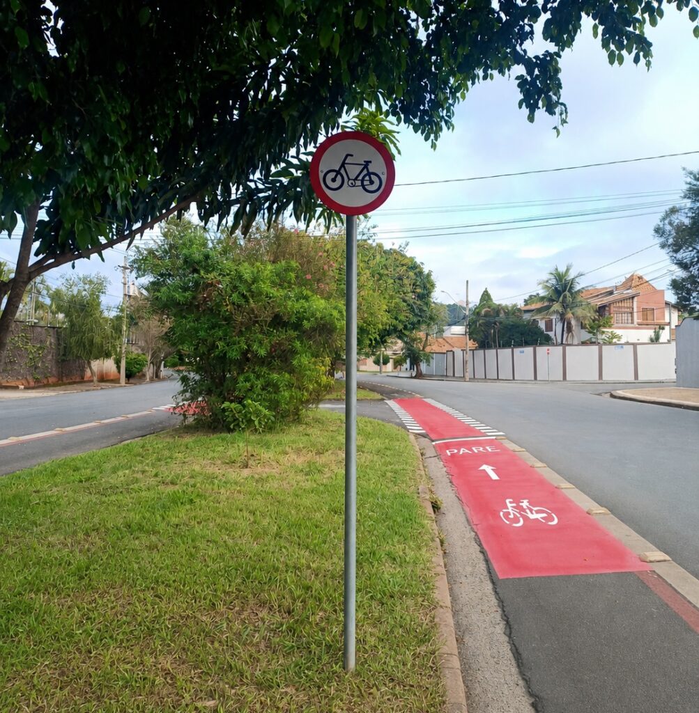 A imagem mostra uma ciclovia pintada de vermelho em uma rua residencial. No centro da imagem, há uma placa de trânsito circular com borda vermelha e um símbolo preto de bicicleta, indicando que o caminho é destinado para ciclistas. No chão, a ciclovia está claramente demarcada com tinta branca, contendo a palavra "PARE", uma seta apontando para frente e o desenho de uma bicicleta, sinalizando que os ciclistas devem parar antes de seguir adiante. Ao redor, há gramado verde, árvores e casas, sugerindo um bairro tranquilo e arborizado. O céu está claro, indicando que a foto foi tirada durante o dia. A imagem transmite uma sensação de organização e segurança para os ciclistas.