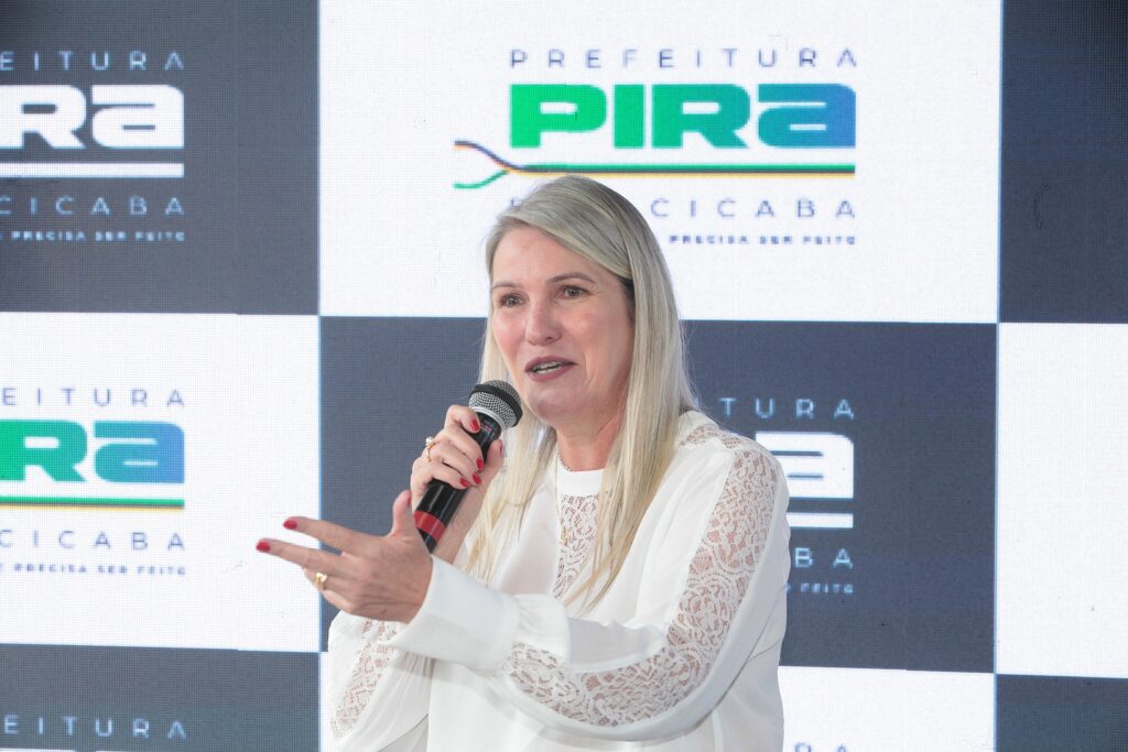 A imagem apresenta uma mulher de cabelos loiros longos e lisos, posicionada no centro, falando ao microfone durante um evento. Ela veste uma blusa branca elegante, com detalhes em renda nas mangas e na parte superior frontal. A mulher mantém uma expressão comunicativa, com a mão esquerda estendida em um gesto de fala, enquanto segura o microfone com a mão direita. Suas unhas estão pintadas de vermelho. Ao fundo, há um painel institucional com um padrão quadriculado em tons de branco e cinza escuro. O painel exibe o logotipo da Prefeitura de Piracicaba, com a palavra "PIRA" em destaque na cor verde, acompanhada pelas cores da bandeira do município (verde, amarelo e azul) em uma linha abaixo, e o slogan "PRECISA SER FEITO" logo abaixo.