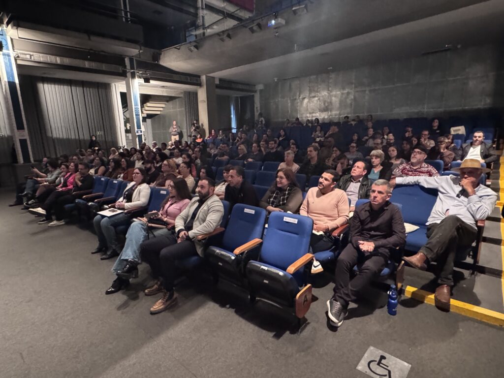 Nesta imagem, vemos um auditório lotado, onde uma plateia está sentada em poltronas azuis, assistindo a uma apresentação ou palestra. O público é variado e parece atento. Em destaque, na parte da frente do auditório: • À esquerda, um grupo de pessoas sentadas, incluindo uma mulher de óculos com um blazer claro e jeans, e homens com camisas sociais e blazers. • À direita, um homem de camisa azul clara, chapéu estilo "panamá" e calça marrom chama a atenção, sentado em uma posição relaxada com a mão no queixo. • Ao lado dele, um homem de camisa preta está sentado com as mãos cruzadas, observando fixamente. O ambiente tem uma estética industrial ou moderna, com paredes de concreto aparente ao fundo e uma estrutura de teto alta. No chão, no canto inferior direito, há uma sinalização de acessibilidade indicando uma vaga ou espaço para cadeirantes.
