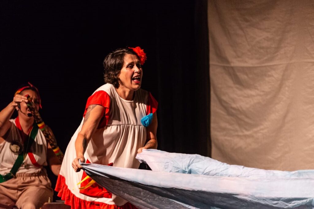 A imagem mostra uma cena teatral com duas mulheres no palco. A mulher em destaque está no centro, vestindo um vestido branco com detalhes vermelhos e uma flor vermelha no cabelo. Ela parece estar cantando ou falando com intensidade, segurando um grande pedaço de tecido azul claro que se espalha à sua frente. Ao lado dela, à esquerda, há outra mulher parcialmente visível, vestindo uma blusa branca com uma faixa colorida (vermelha, verde e amarela) e segurando um objeto que parece ser um instrumento ou adereço cênico. O fundo é composto por um cenário simples, com um tecido claro que serve de cortina ou fundo, e a iluminação está focada nas atrizes, destacando suas expressões e roupas, enquanto o restante do palco está mais escuro. A cena transmite emoção e movimento, sugerindo um momento dramático ou musical da peça.