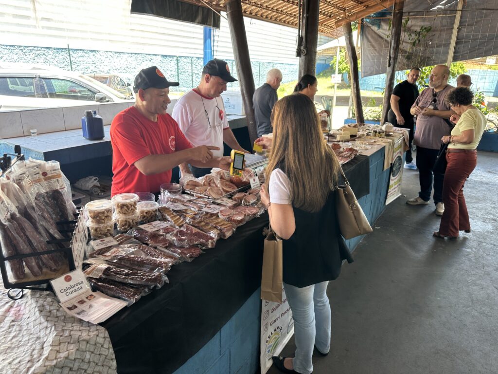 A imagem mostra uma cena em um mercado ou feira ao ar livre, onde uma cliente está realizando uma compra em uma banca de embutidos e defumados. * No centro da imagem, um vendedor usando uma camiseta vermelha e boné está segurando uma máquina de cartão, apontando para ela enquanto interage com uma cliente. Ao lado dele, outro vendedor de camiseta branca observa a transação. * A banca está repleta de produtos embalados a vácuo, incluindo linguiças, carnes curadas e potes com torresmos. Há etiquetas de identificação nos produtos, como uma que diz "Calabresa Curada" em primeiro plano. * A cliente, de costas para a câmera, veste uma blusa branca e colete preto, segurando uma sacola de papel. Ela está prestes a realizar o pagamento. * Ao fundo, é possível ver outras pessoas circulando pela feira, algumas olhando outros produtos da banca, que também oferece queijos expostos mais ao fundo. O ambiente parece ser uma estrutura coberta, com colunas de madeira e piso de concreto.