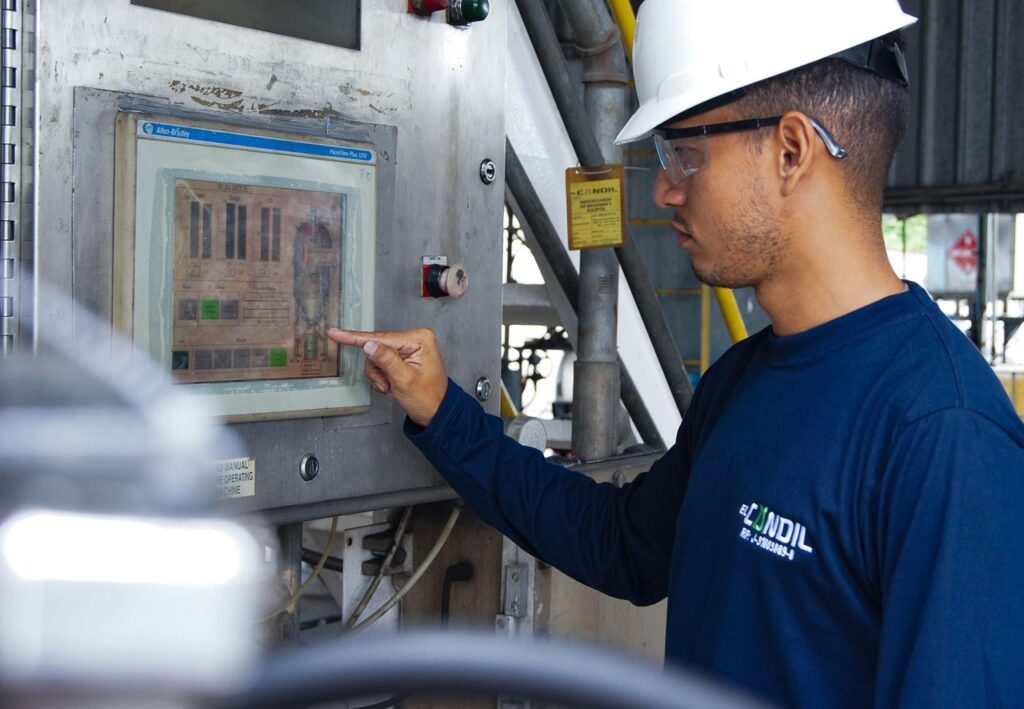 A imagem mostra um homem de perfil, trabalhando em um ambiente industrial. Ele usa um capacete branco de segurança, óculos de proteção e uma camiseta azul escura com o logo "EL CANDIL" no peito. O homem está operando um painel de controle industrial da marca Allen-Bradley, modelo PanelView Plus 1250. Ele aponta com o dedo indicador para a tela do painel, que exibe gráficos de barras e um esquema de um tanque, provavelmente relacionado a processos de controle industrial, com indicações como "Vapor", "Lodo" e "Óleo". Ao fundo, há tubulações metálicas, estruturas de metal e uma etiqueta amarela pendurada em um tubo, indicando um ambiente típico de fábrica ou instalação industrial. A iluminação é clara, sugerindo que o local seja bem iluminado, possivelmente com luz natural. A imagem transmite uma atmosfera profissional, focada em monitoramento e operação de equipamentos industriais.
