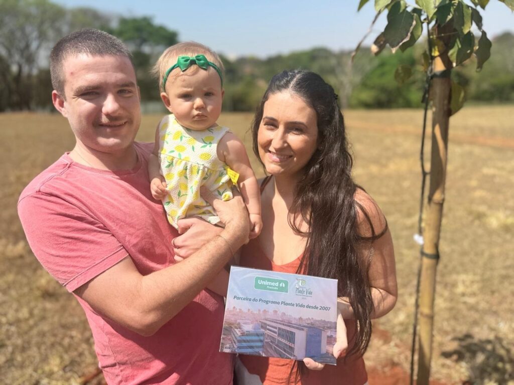 A imagem mostra uma família composta por um homem, uma mulher e um bebê, posando ao ar livre em um campo durante o dia. O homem, à esquerda, veste uma camiseta cor de salmão e segura o bebê, que está vestido com um vestido amarelo estampado com limões e usa uma faixa verde na cabeça. A mulher, à direita, veste uma blusa regata laranja e segura uma placa comemorativa, que exibe os logos da Unimed Piracicaba e do programa Plante Vida, além de uma foto de um prédio hospitalar. Ao lado deles, há uma muda de árvore jovem, amparada por uma estaca de bambu. O cenário é um campo com grama seca sob um céu azul claro, indicando um dia ensolarado. A imagem transmite uma mensagem de envolvimento com a preservação ambiental e responsabilidade social, destacando a parceria da Unimed Piracicaba com o programa Plante Vida desde 2007.